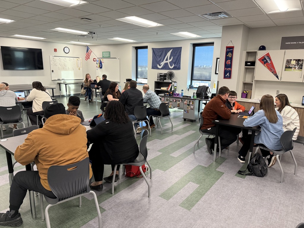 Students and parents sit at desks in a classroom chatting with teachers. 