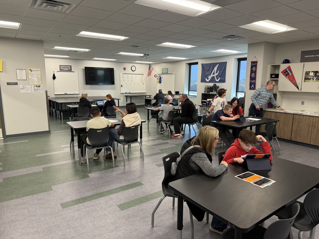 Students and parents sit at desks in a classroom chatting with teachers. 