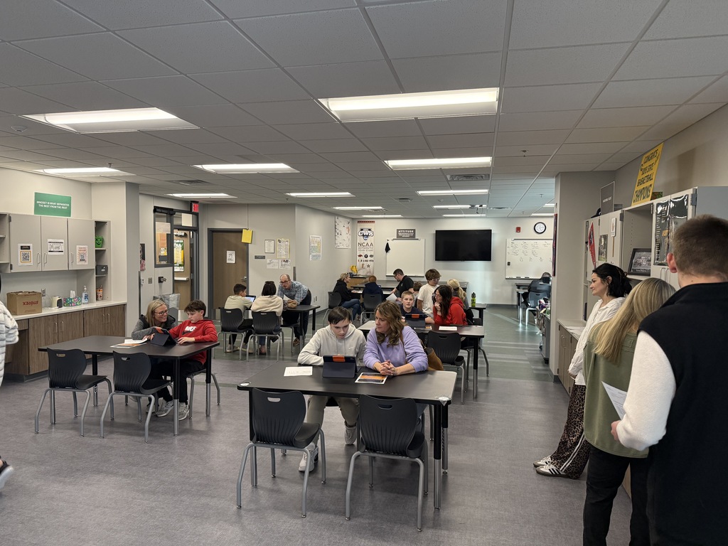Students and parents sit at desks in a classroom chatting with teachers. 