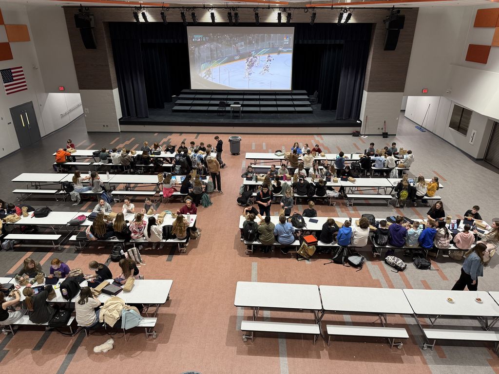 Image from above a cafeteria, looking down on students seated at cafeteria lunch tables. 