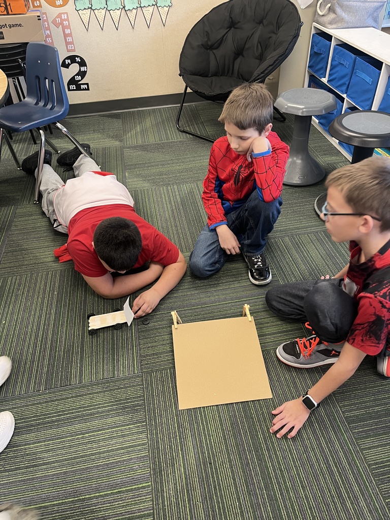 Three boys sitting in a classroom on a ground looking at a car made out of popsicle sticks and a cardboard ramp on the ground