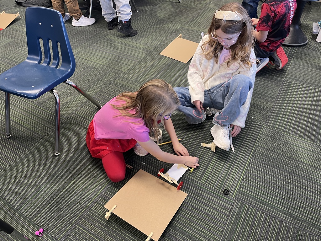 Two girls working on a carpet in a classroom on an activity with a paper car on a cardboard ramp
