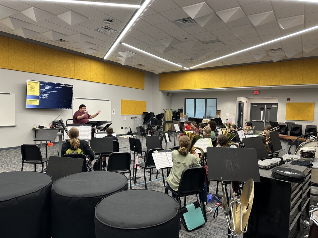 A band teacher sits in front of the room conducting members of a student band. 