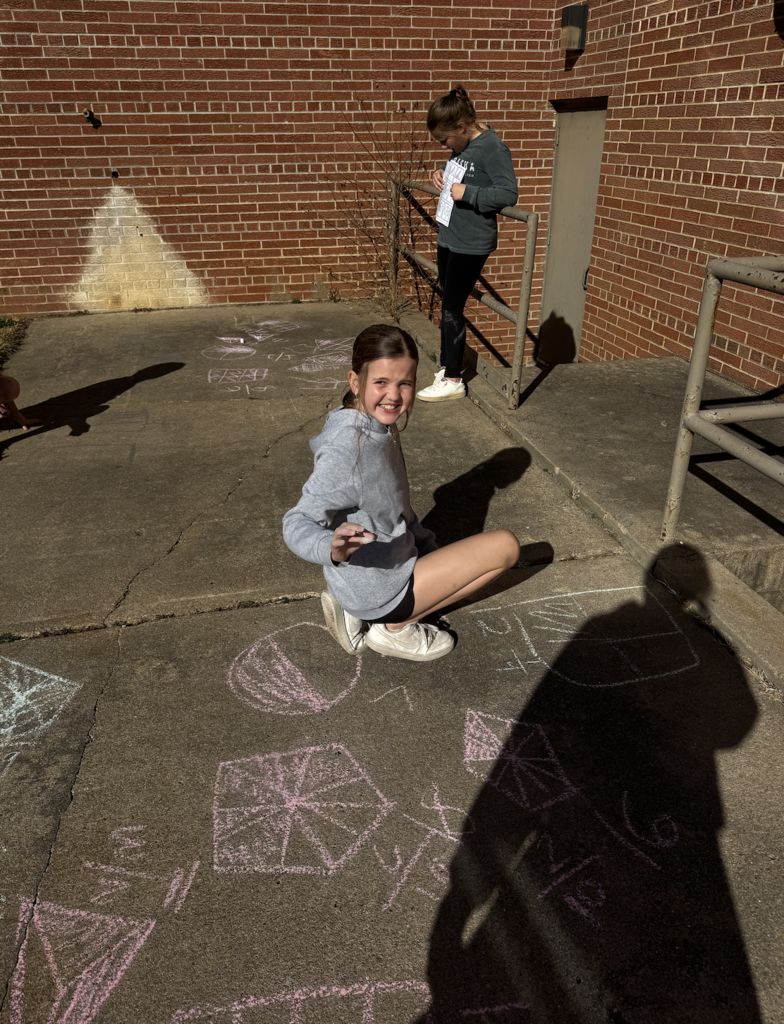 Girl kneels on the ground smiling while chalk drawings show on the cement.