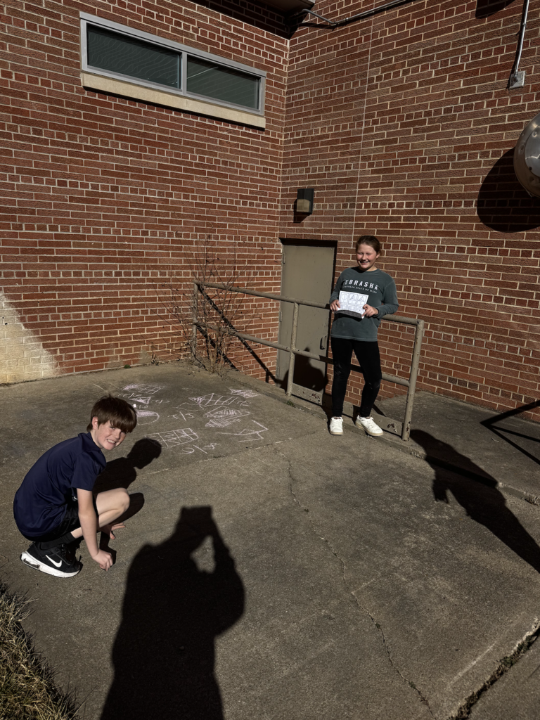 Girl stands smiling holding a paper while a boy kneels on cement with chalk drawings present.