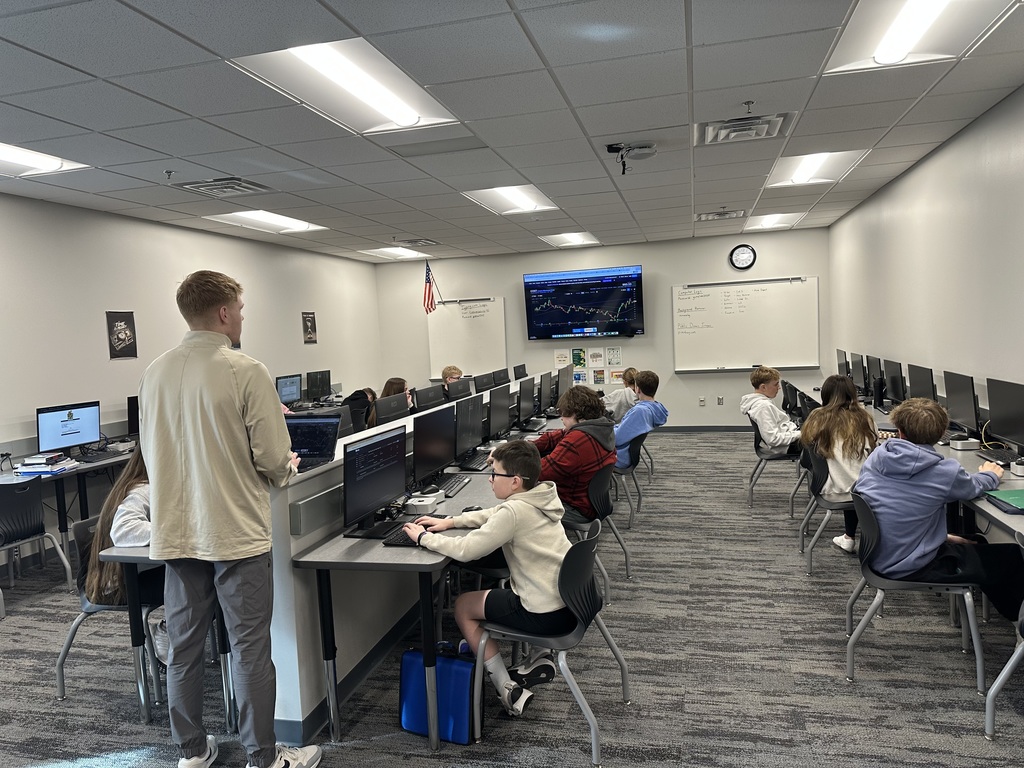 Students sit at computers around a room with a teacher in the back. There is a screen in the front with stock market graphics. 