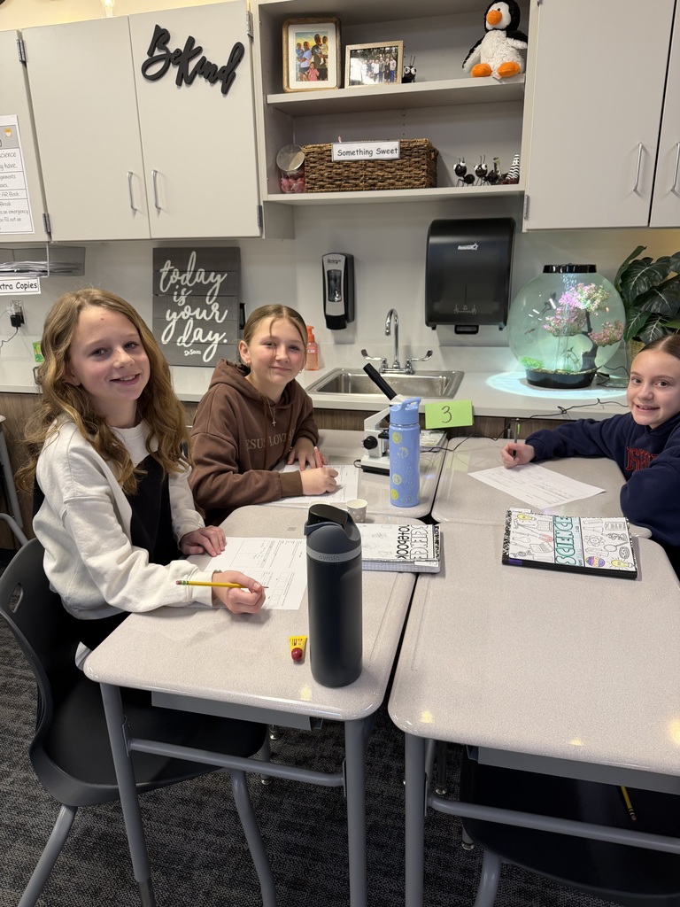3 girls sit at desks, smiling at the camera. 