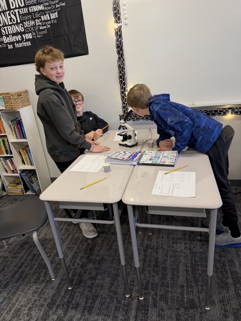 3 boys are at desks. One is looking in a microscope while the other two look at the camera. 
