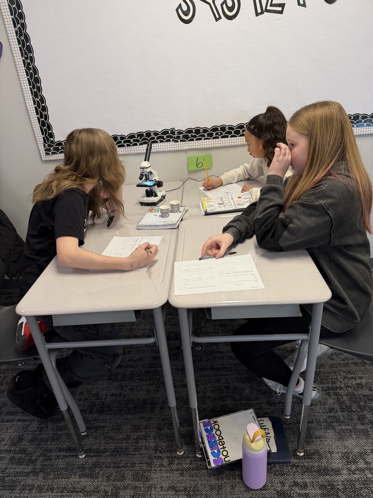 3 girls sit at desks, looking at microscopes and lenses.