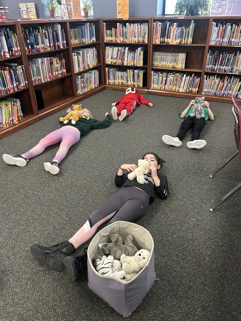 Four students lie down on carpet in a library with a stuffed animal on them.