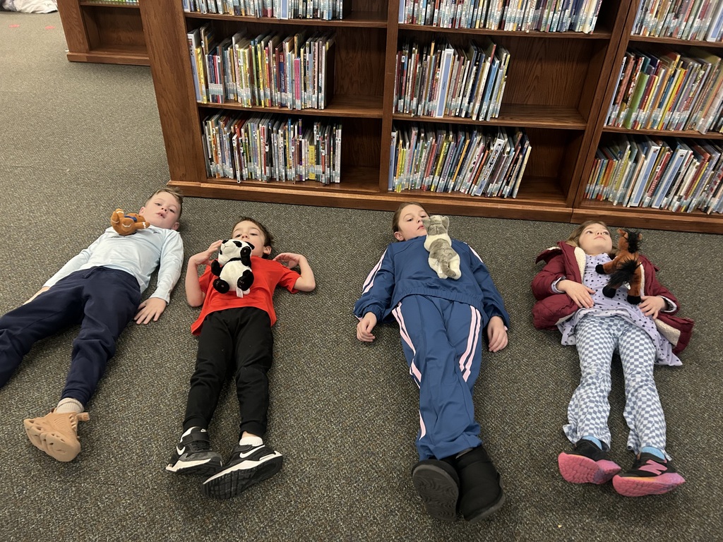 Four students lie down on carpet in a library with a stuffed animal on them.