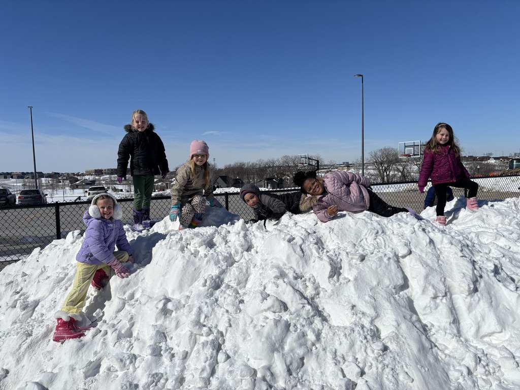 6 first graders stand atop a snow pile smiling.