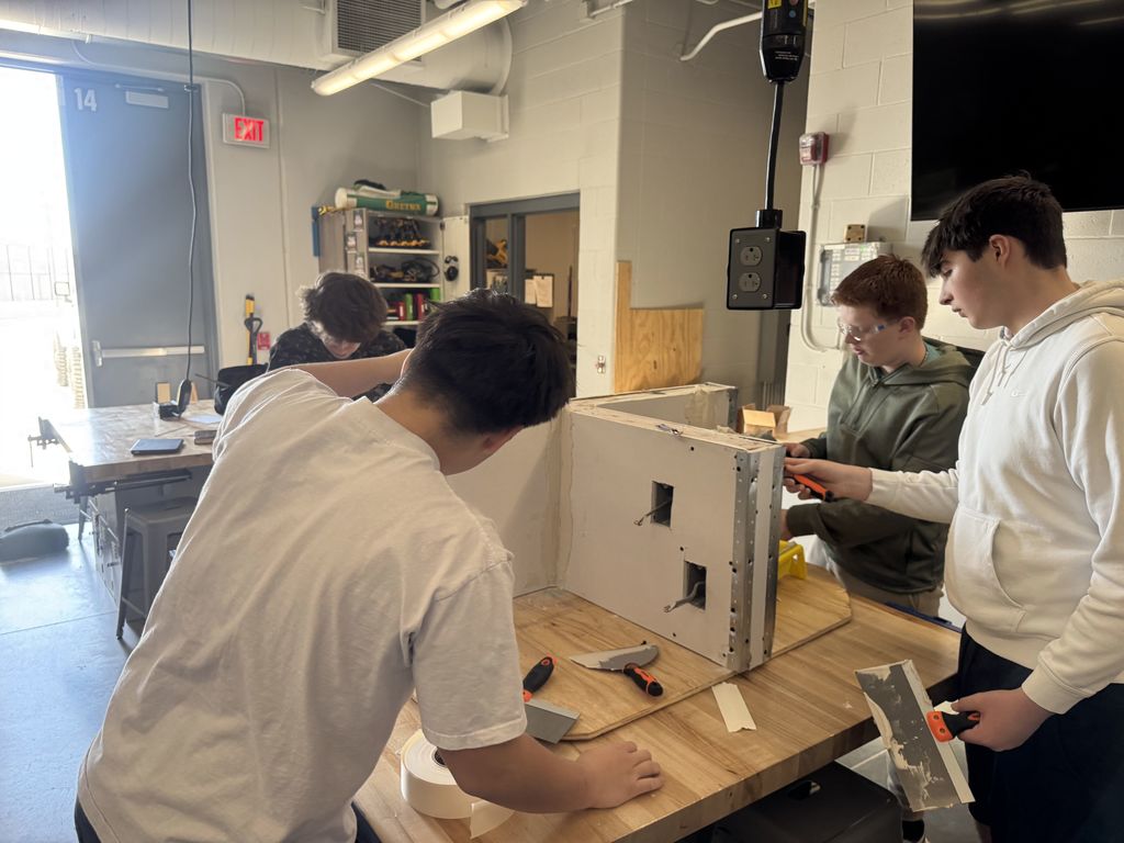 Four students stand around a table working on a drywall project. 
