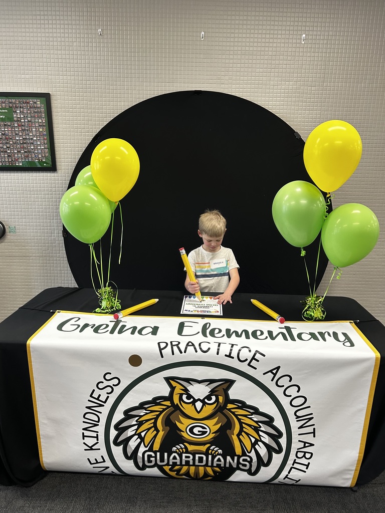 A young boy in a striped shirt sitting at a "Gretna Elementary Guardians" table, signing a document with a giant yellow novelty pencil.