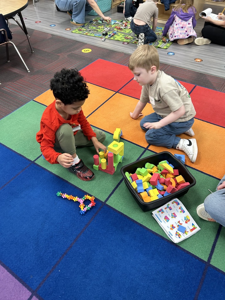 Two young boys sitting on a colorful checkered rug, playing with building blocks and vibrant plastic flower-shaped connectors.