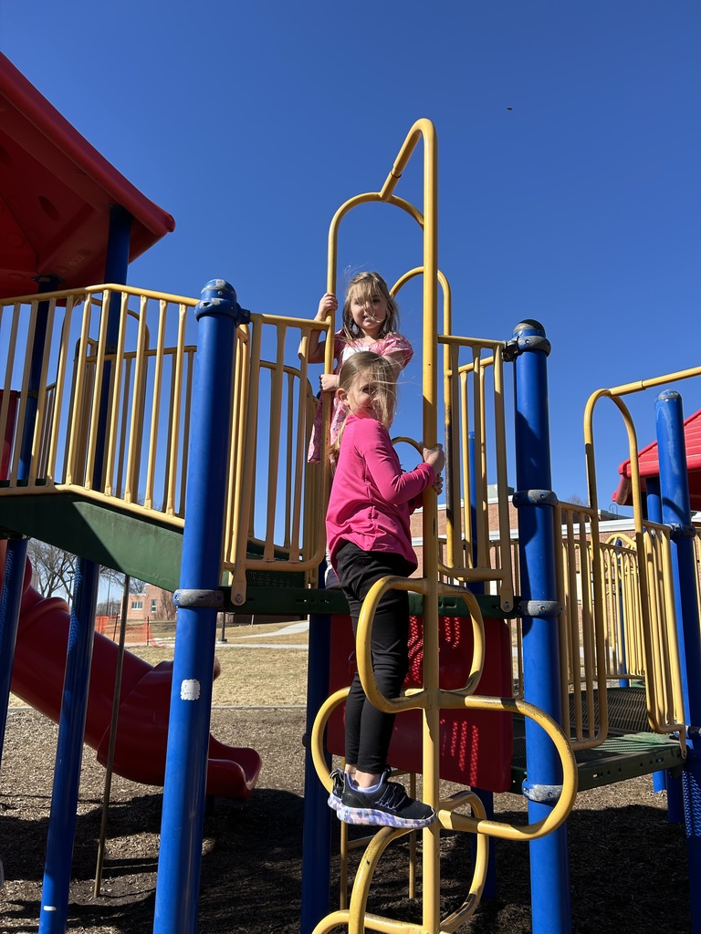 Two young girls smiling while climbing on a yellow and blue playground structure under a clear blue sky.