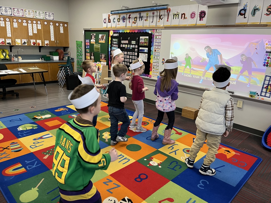 Children wearing white paper headbands standing on a colorful alphabet rug while following an interactive dance video on a large classroom screen.