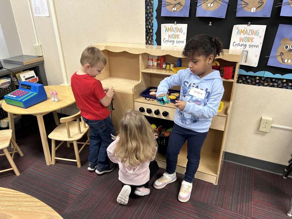 Three young children engaged in imaginative play within a classroom play kitchen area featuring wooden shelves and toy food.
