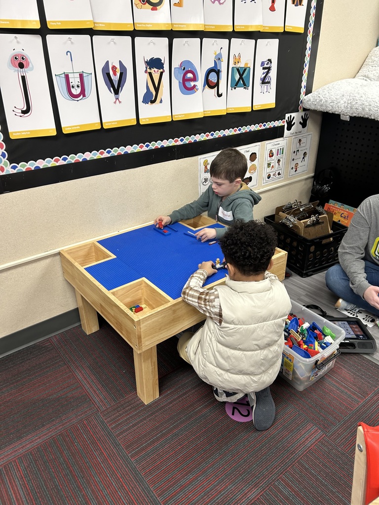 Two children sitting at a wooden Lego table with a blue baseplate, building structures with colorful bricks.