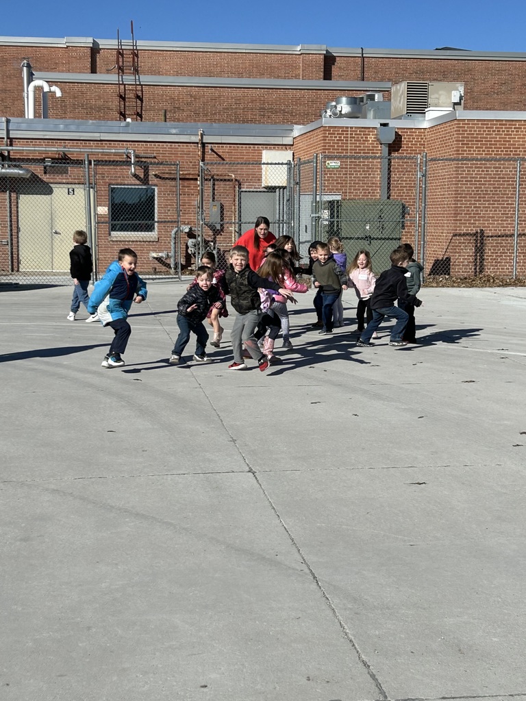 A group of elementary students running and playing across a concrete playground area on a bright, sunny day.