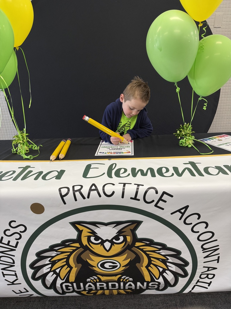 A young boy in a blue jacket focused on signing a paper with a giant novelty pencil at a decorated Gretna Elementary event table.