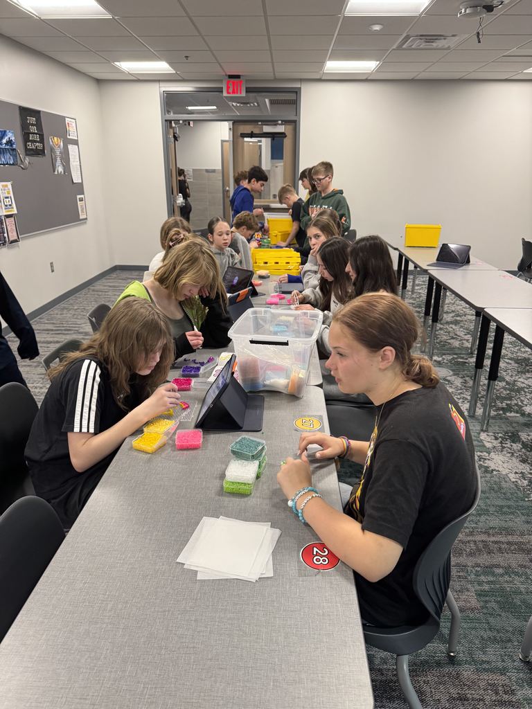 Students sit at a long table working with Legos and Perler Beads. 