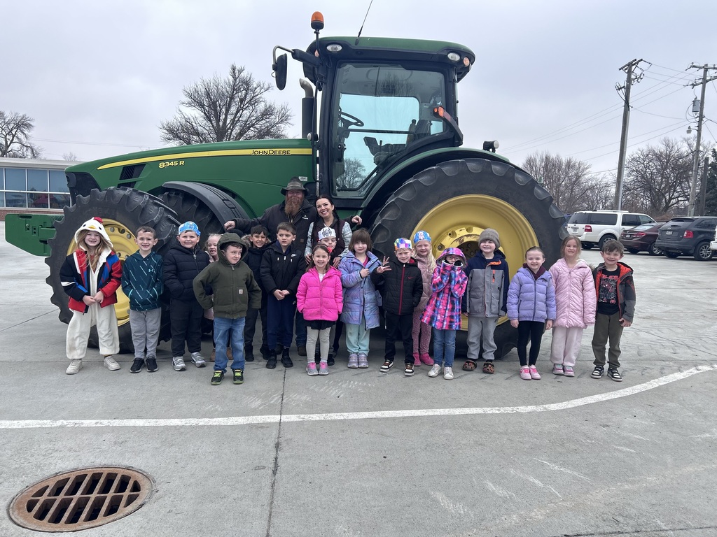 A group of children posing with two adults in front of a large green John Deere tractor on a gray, overcast day.