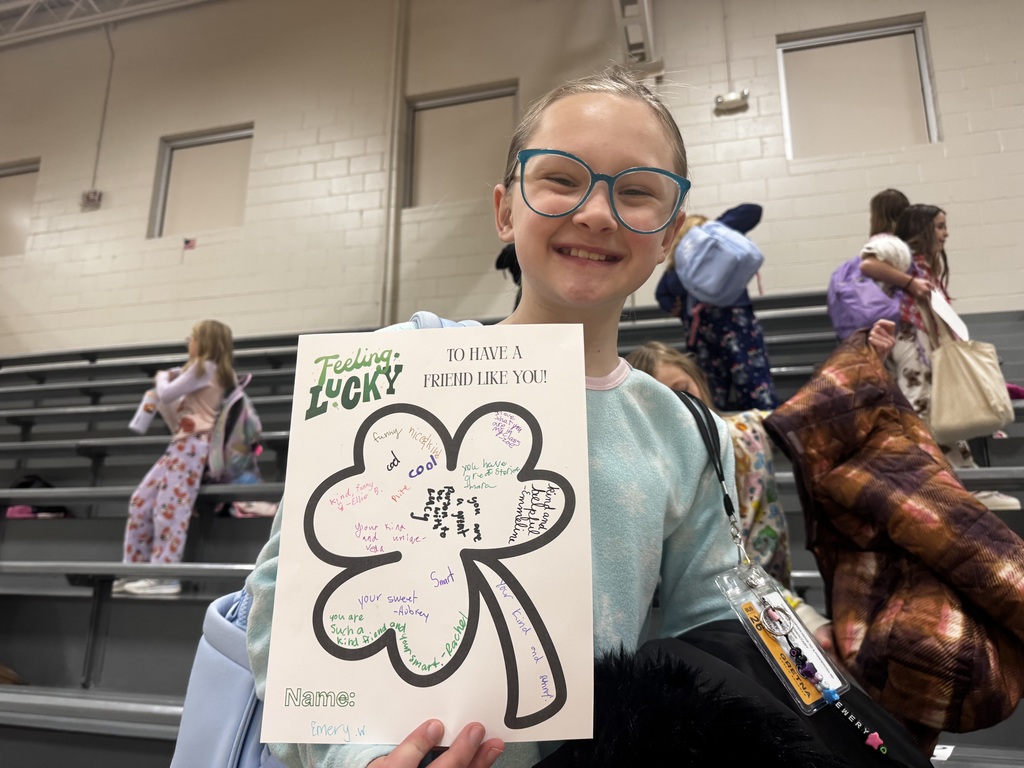 A close-up of a smiling girl with blue glasses holding her clover sheet. Her name, "Emery W.," is at the bottom. The clover contains sweet messages like "Kind and helpful," "You have great stories," and "You are a great person to be with." She is wearing a light blue snowflake-patterned top.