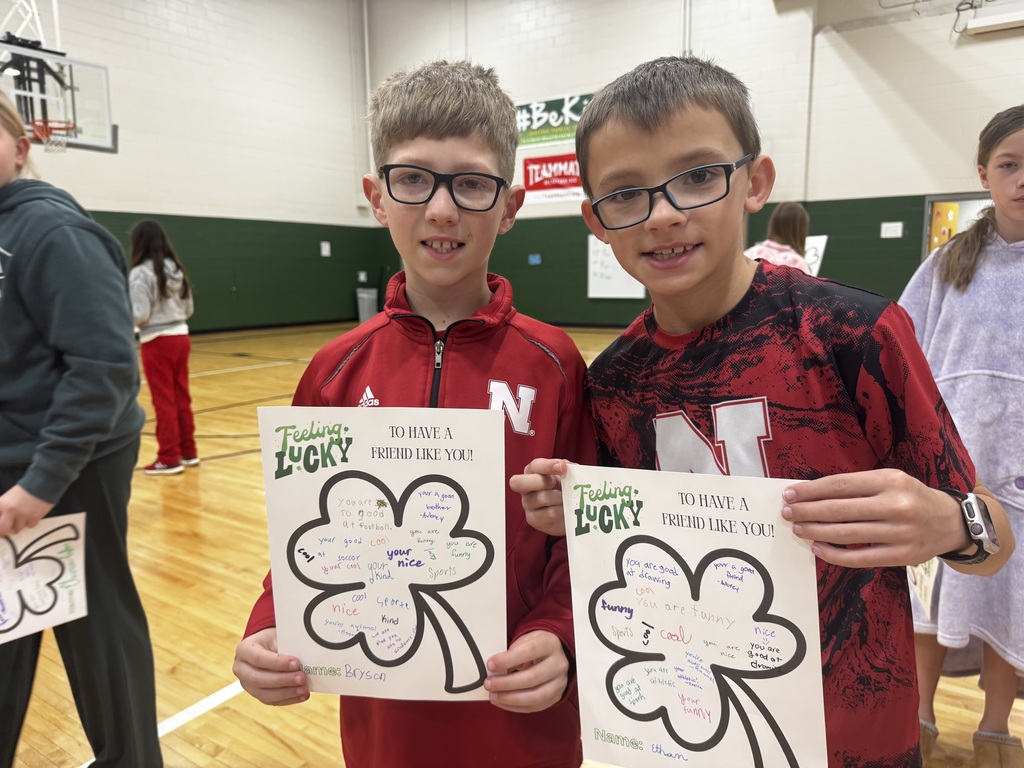 A medium shot of two young boys standing in a gym, wearing glasses and athletic shirts. They are proudly holding up their "Feeling Lucky" clover papers. The clovers are filled with handwritten compliments from their peers, such as "You are good at football" and "You are funny." Their names, "Bryson" and "Ethan," are written at the bottom of their respective sheets.