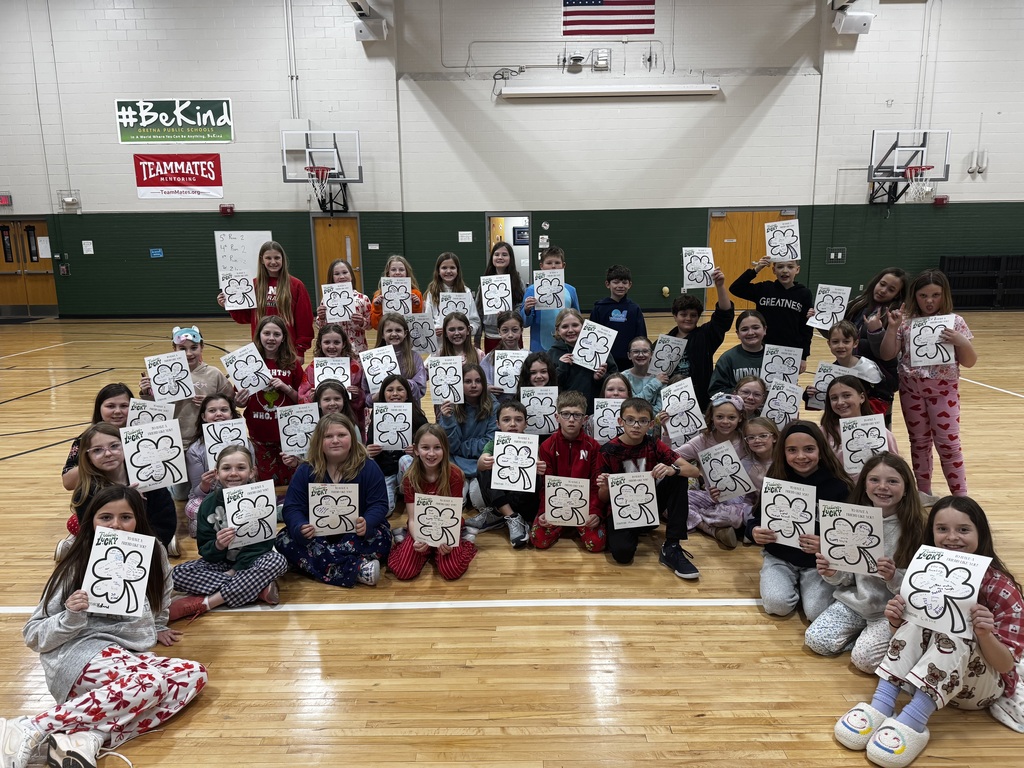 A wide shot of about 40 elementary-aged children sitting and standing in a school gymnasium. Every child is holding up a white sheet of paper featuring a large, hand-drawn four-leaf clover. Above them on the wall are signs that read "#BeKind" and "Teammates Mentoring." The children are dressed in casual clothes and pajamas, smiling for the camera.