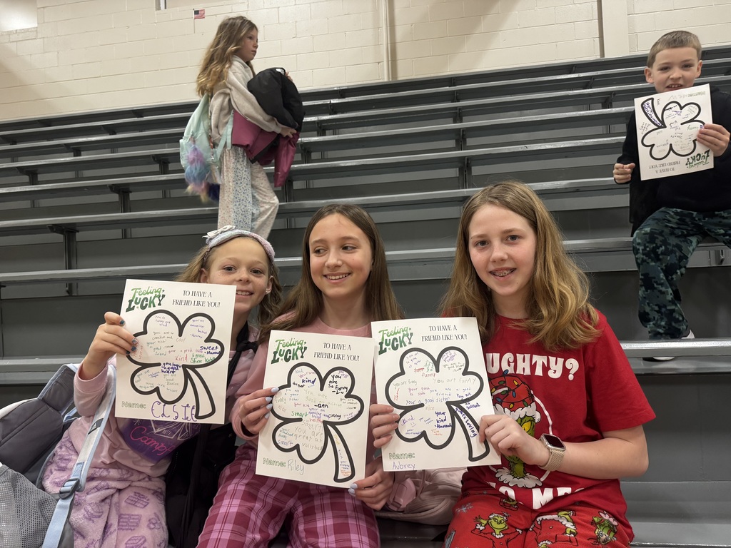 Three girls sit together on gray bleachers, each holding their decorated clover paper. They are smiling warmly at the camera. From left to right, their names are Elsie, Riley, and Aubrey. The girl on the far right wears a red "Grinch" themed shirt. The papers serve as a collection of positive affirmations from their classmates.