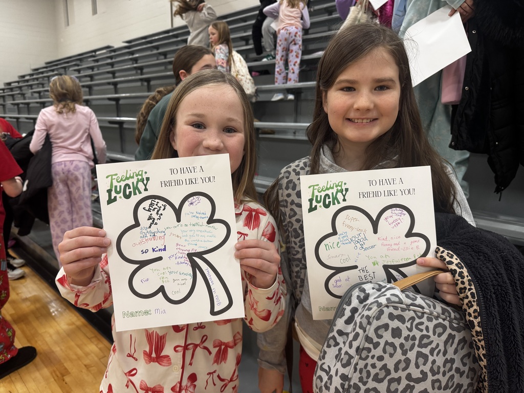 Two girls smile while holding their "Feeling Lucky" papers. The girl on the left, Mia, is wearing red bow-patterned pajamas. The clovers are densely packed with messages in various ink colors. In the background, other students are visible on gray gym bleachers.