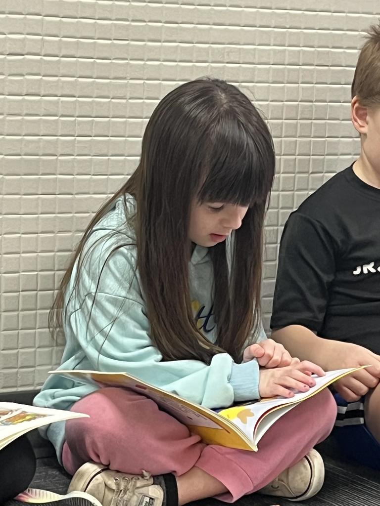 A young girl with long brown hair wearing a light blue hoodie, sitting cross-legged and intently reading a picture book.