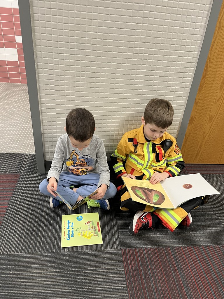 Two young boys sitting cross-legged on a carpeted floor reading books; one boy is wearing a yellow firefighter costume.