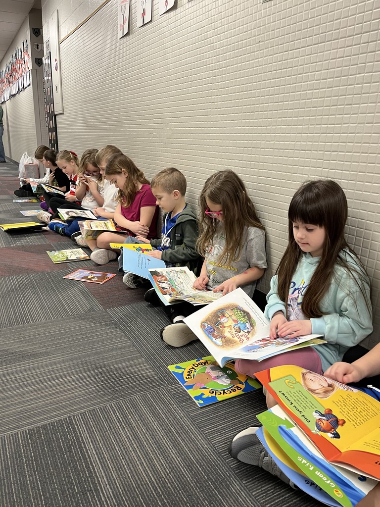 A long line of young children sitting on a hallway floor, leaning against a tiled wall while focused on reading various colorful picture books.