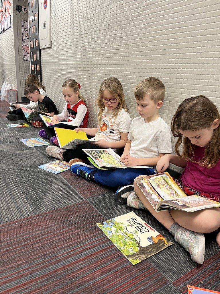 A side view of several children sitting in a row against a school hallway wall, each deeply engaged in reading their own illustrated book.