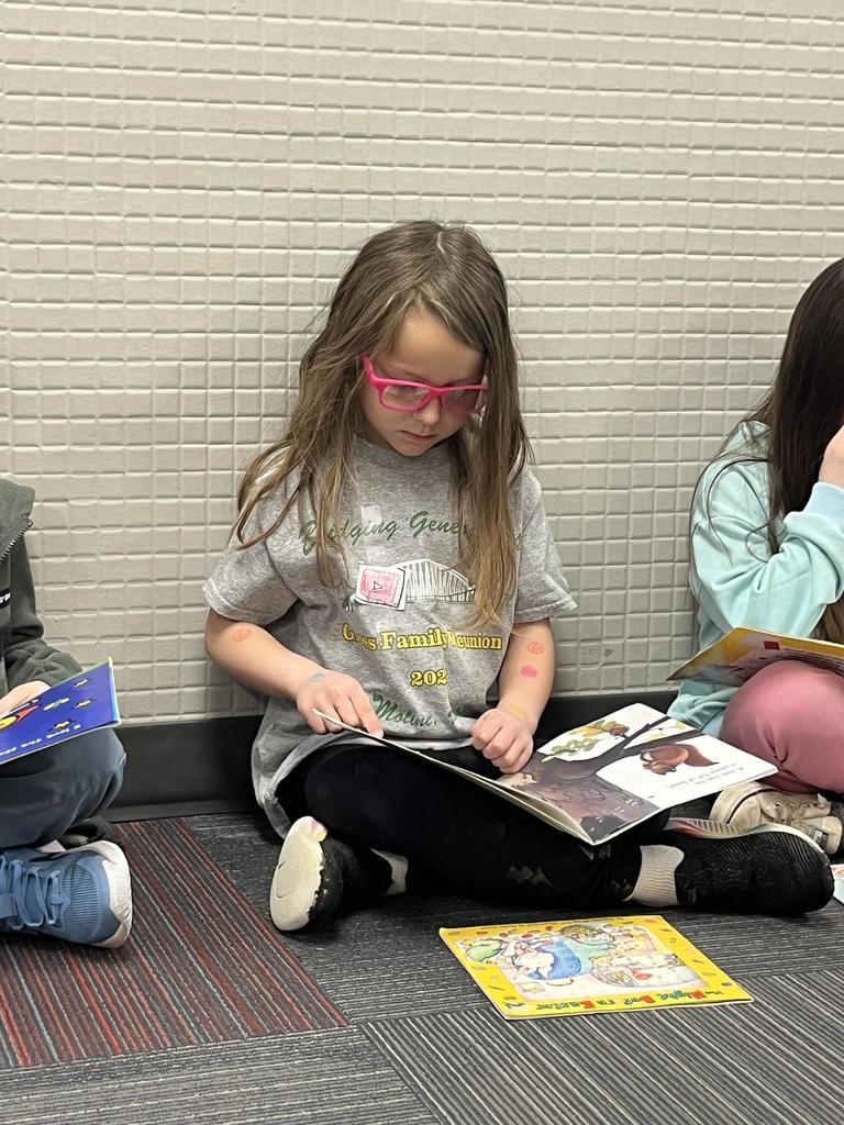 A young girl with pink glasses sitting on a gray and red carpet, looking down and turning the pages of an illustrated storybook.