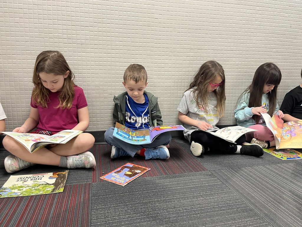 Four children sitting side-by-side against a neutral tiled wall, showing a variety of colorful books spread out on their laps and the floor.
