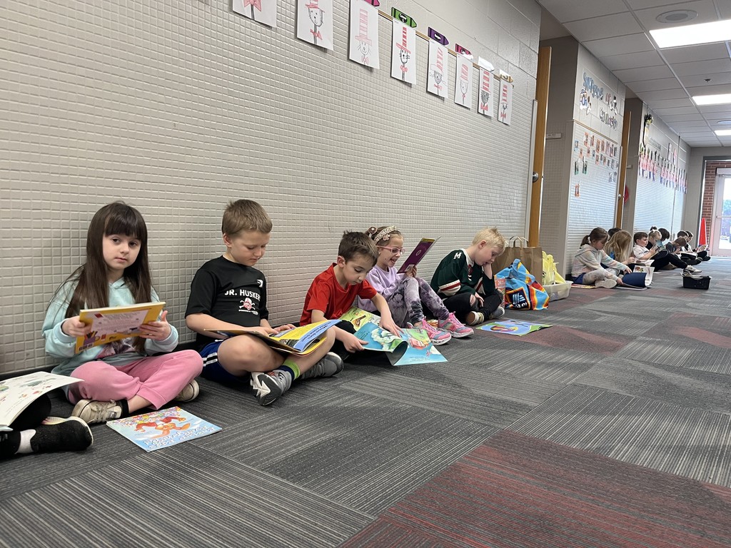A wide shot of a school hallway where a line of children are sitting and reading under a display of "Cat in the Hat" drawings.