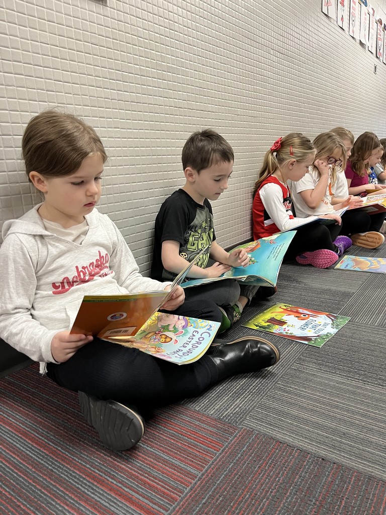 Close-up of three children sitting on a patterned carpet reading; the girl in front reads a "Corduroy" book while the others look at theirs.