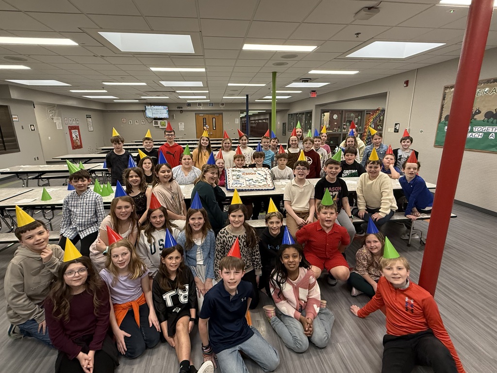 A large group of elementary-aged children wearing colorful party hats posing behind a long table in a school cafeteria with the Nebraska-themed cake.