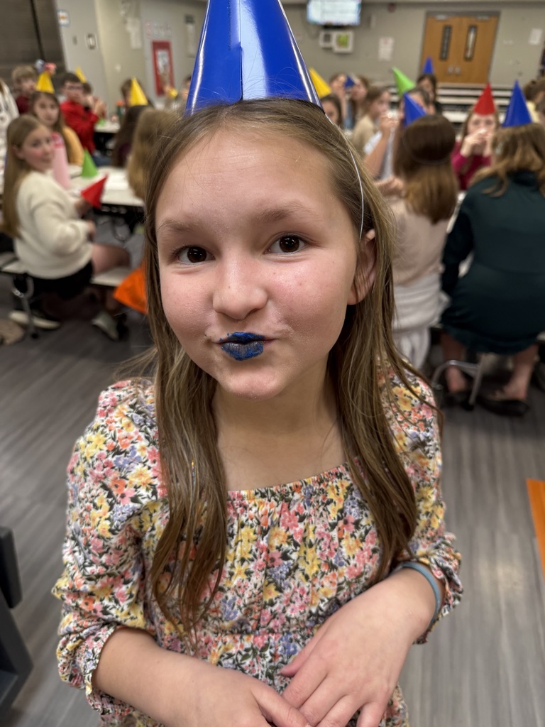 A close-up of a young girl in a floral dress and blue party hat, playfully showing off her blue-stained lips from the cake frosting.