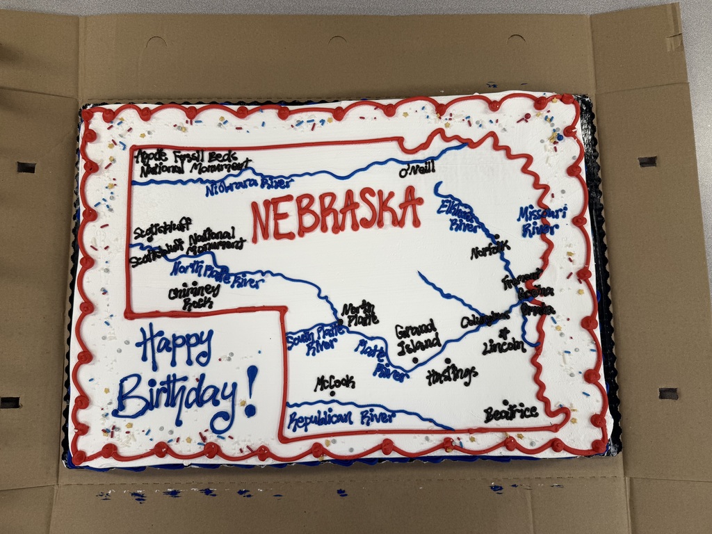 A rectangular sheet cake decorated with a hand-drawn map of Nebraska in frosting. It features blue rivers, black labels for major cities and landmarks, and "Happy Birthday!" written in the corner.
