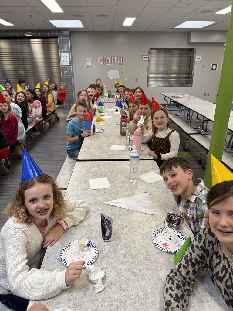 A line of children sitting at a cafeteria table wearing party hats, eating cake, and drinking juice pouches.