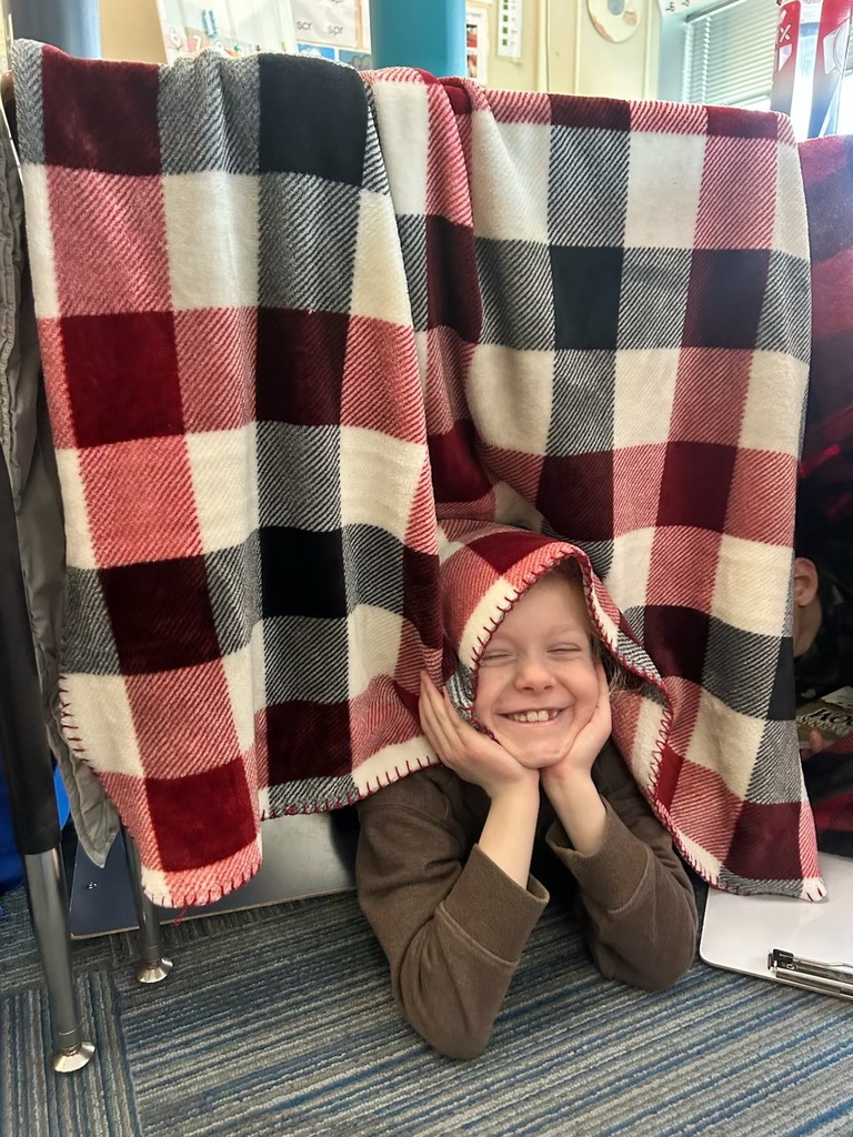 A young child lies on the floor, smiling with their head resting on their hands, framed by red, white, and black plaid blankets draped over a desk.