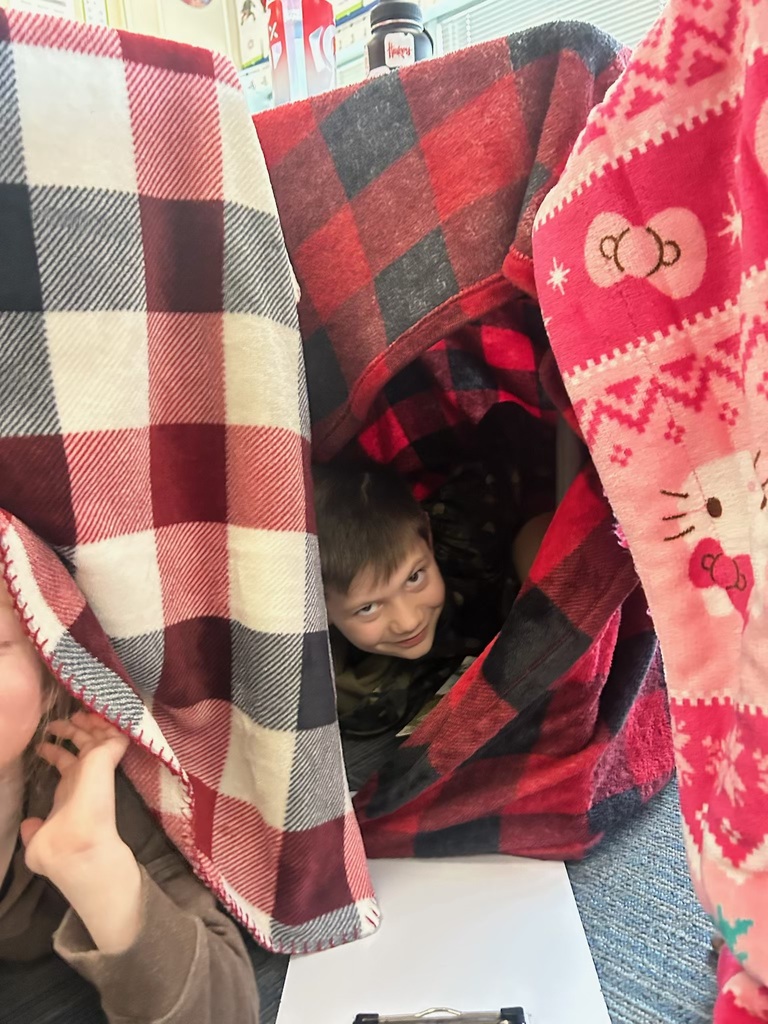 A boy peeks out from a gap between a red and black plaid blanket and a pink Hello Kitty blanket used to create a fort over a classroom desk.