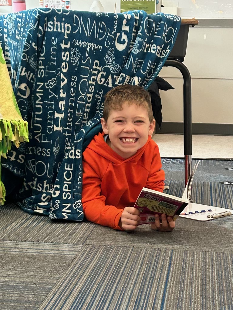 A boy in a bright orange hoodie grins broadly while lying on a classroom floor, holding a book next to a desk draped with a blue blanket featuring printed words.