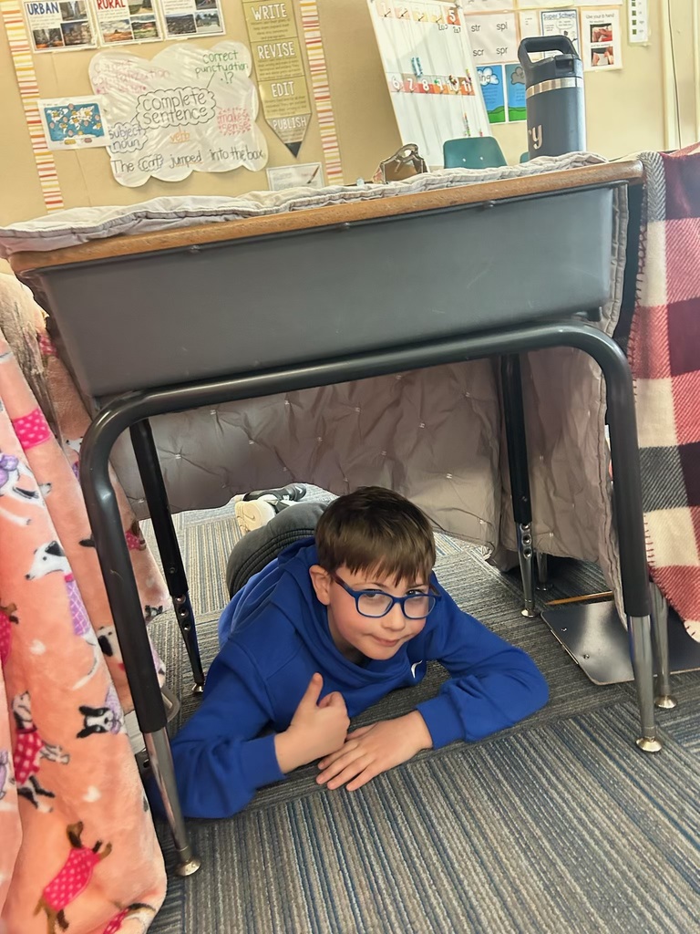 A boy wearing blue-rimmed glasses and a blue hoodie gives a thumbs-up while lying under a desk covered with a grey quilted blanket in a classroom.