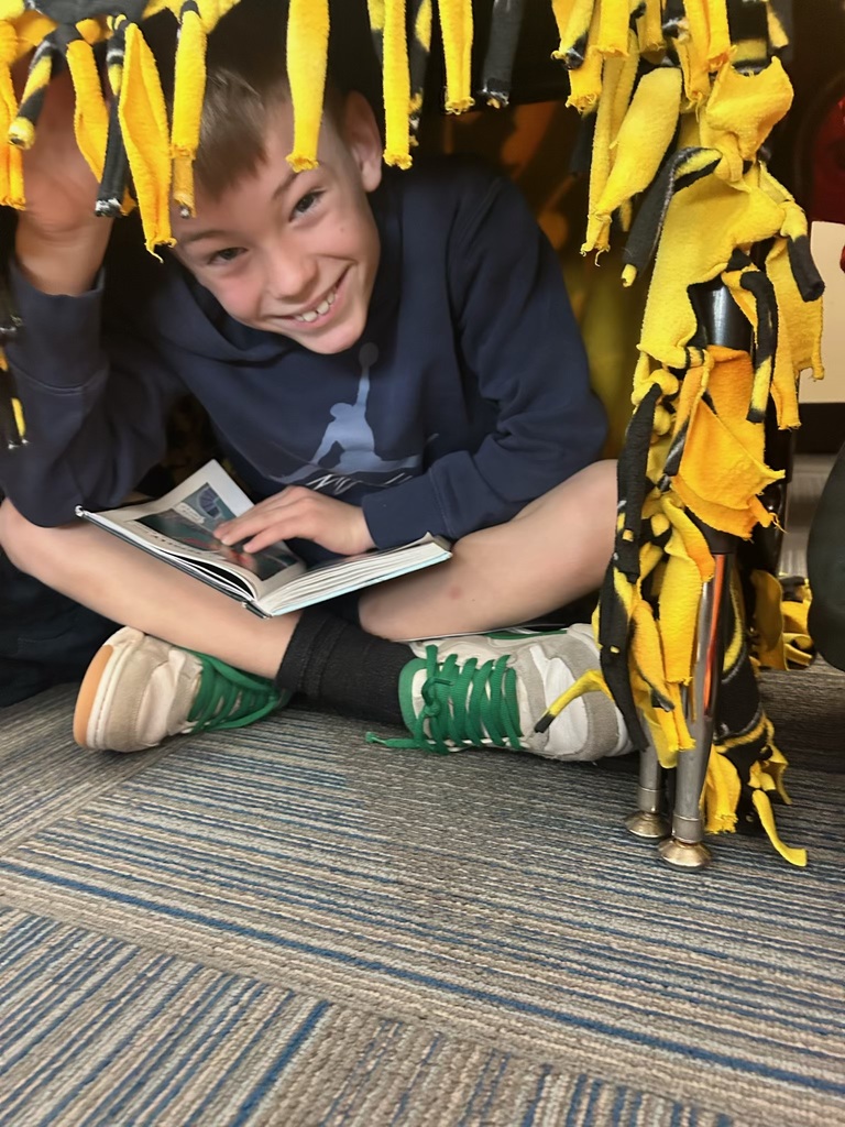 A smiling boy in a dark blue hoodie sits cross-legged under a desk adorned with yellow and black fringed fabric, holding an open book.