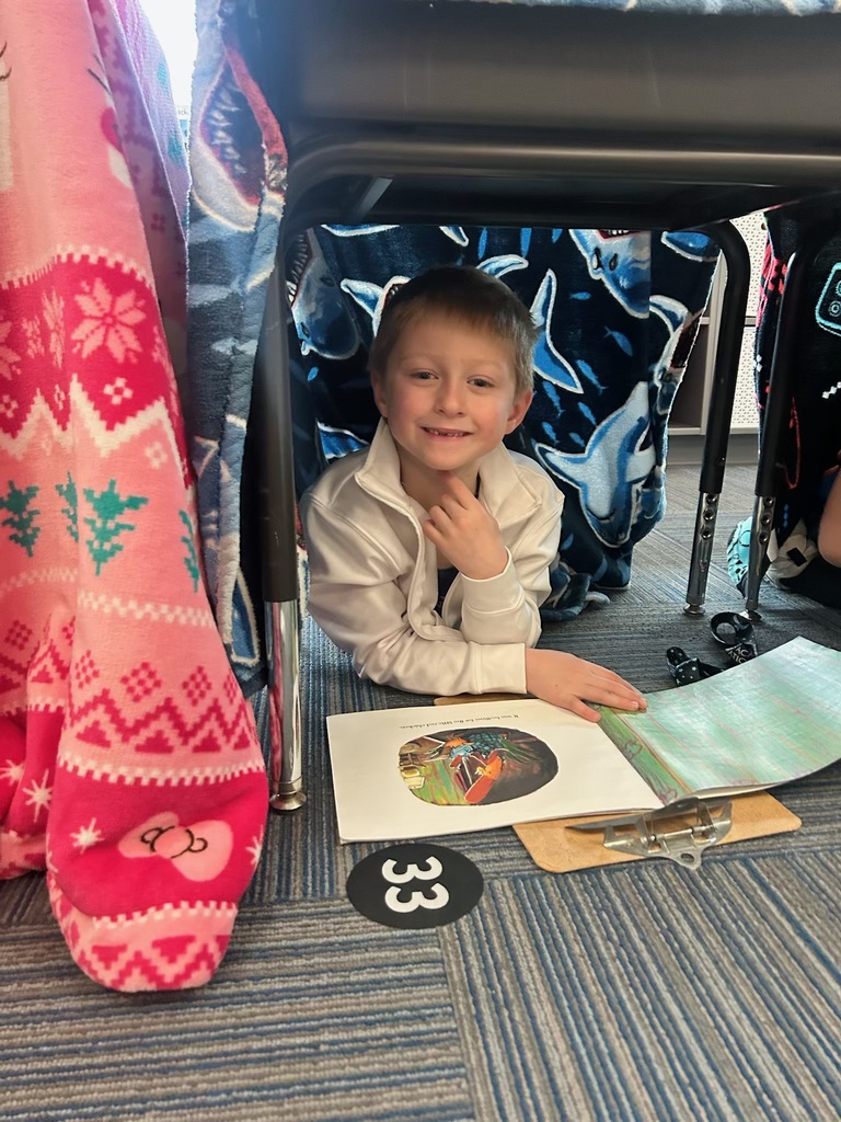 A young boy in a white jacket smiles while lying on a classroom floor inside a fort made of shark-patterned and pink blankets draped over desks. He has an open book and a clipboard in front of him.